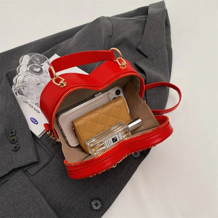 Red heart-shaped handbag with gold zipper and handle on gray blazer, containing smartphone, quilted wallet, and perfume bottle