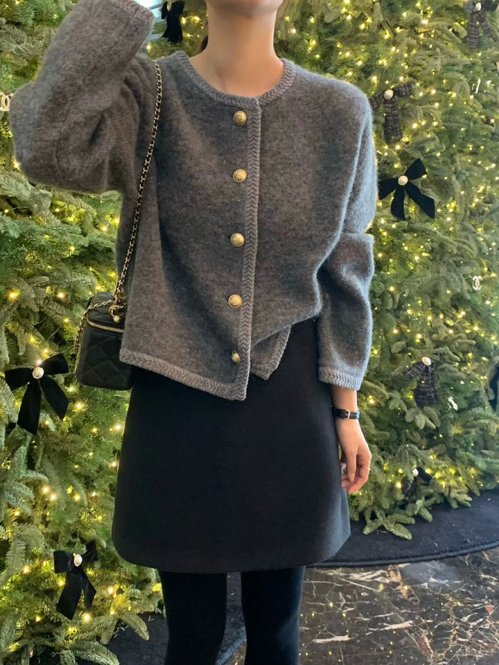 Woman wearing gray buttoned cardigan and black skirt standing by decorated Christmas tree with lights and bows