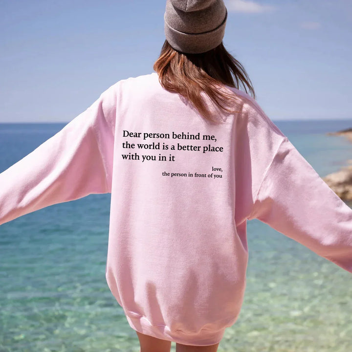 Person wearing pink sweatshirt with motivational text by the sea, sunny day, wearing brown beanie