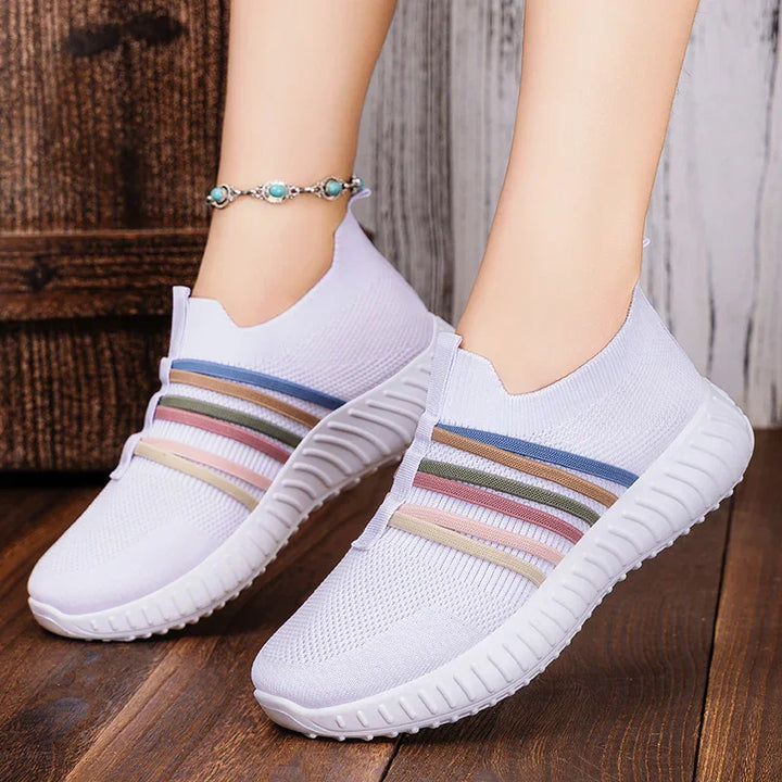White slip-on sneakers with multicolor elastic bands worn on feet against wooden floor