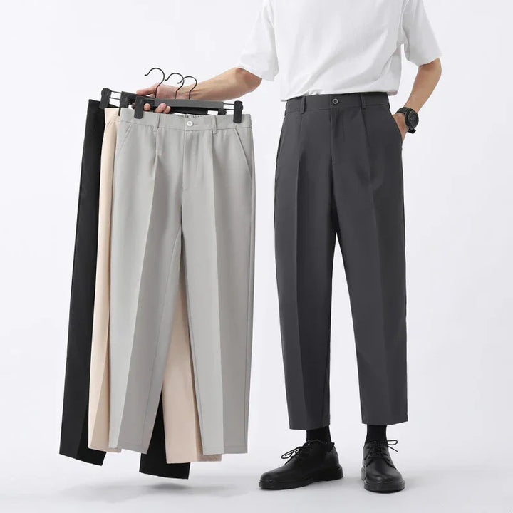 Man wearing dark gray cropped trousers and black shoes holding hangers with black, beige, and gray pants