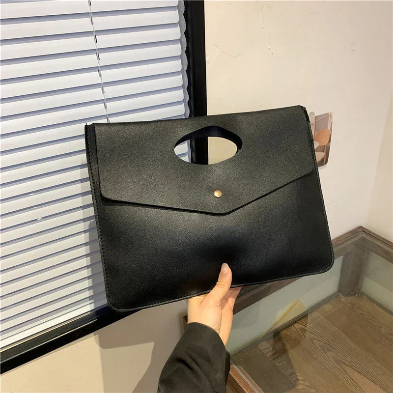 Hand holding a sleek black leather clutch bag with a top oval handle and gold button clasp near window blinds
