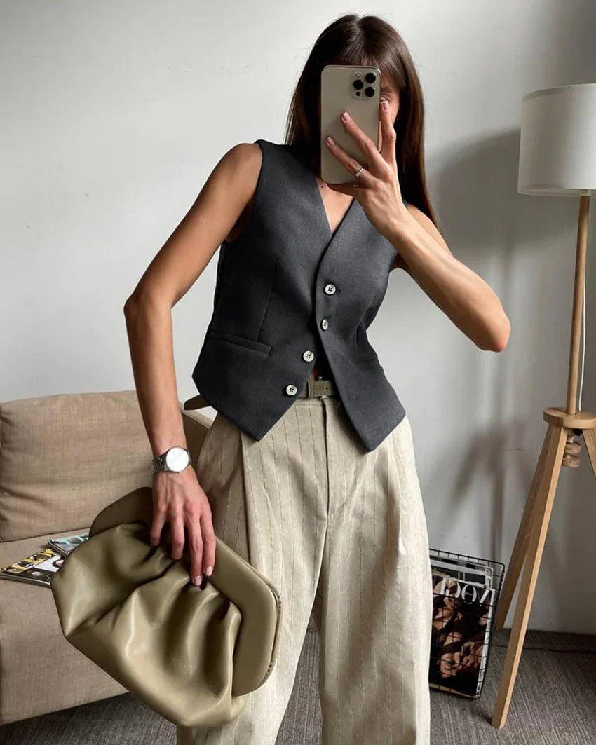 Woman wearing dark gray buttoned vest and beige striped wide-leg pants holding olive green clutch in modern living room
