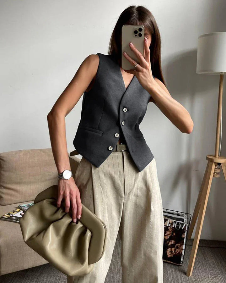 Woman wearing dark gray buttoned vest and beige striped wide-leg pants holding olive green clutch in modern living room