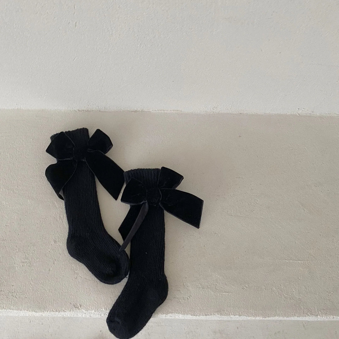 Pair of black knit knee-high socks with large velvet bows on a beige textured floor