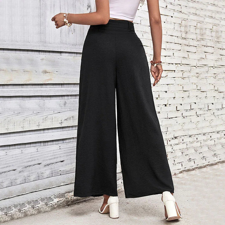 Woman wearing high-waisted black wide-leg pants with white block heel sandals against a brick wall