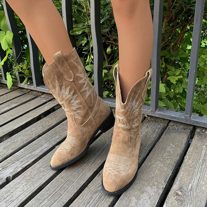 Close-up of brown suede cowboy boots with white stitching worn on wooden deck near metal railing and green foliage
