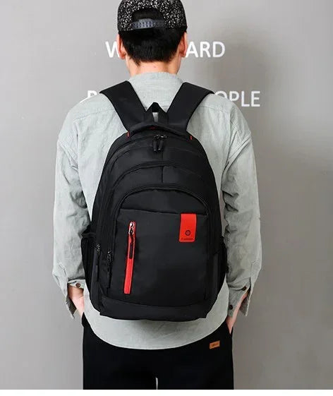 Man wearing black backpack with red accents and multiple zippered compartments against gray wall