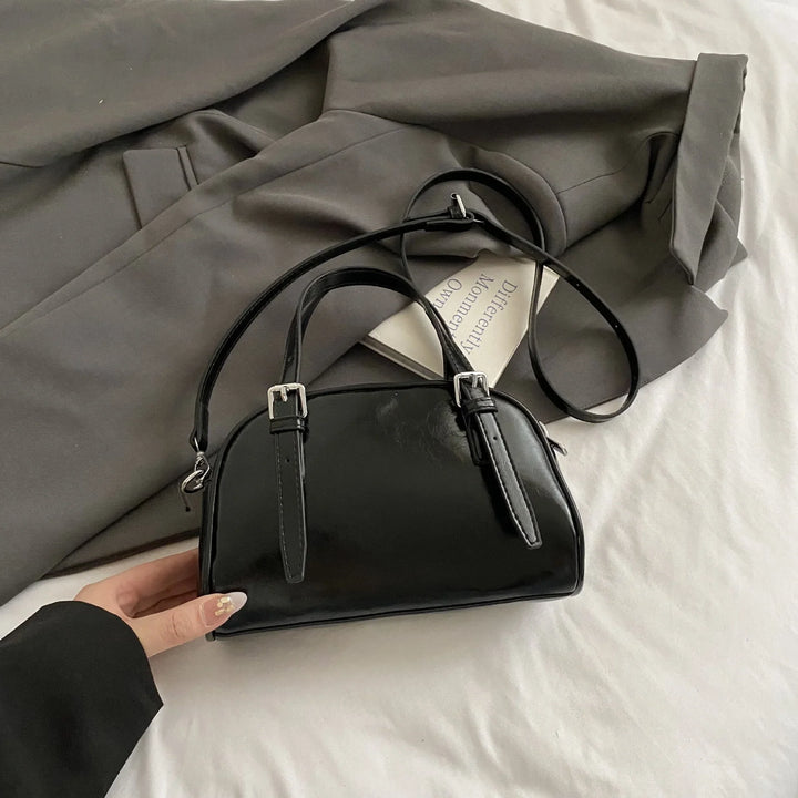 Glossy black leather handbag with adjustable straps on gray jacket and white bed