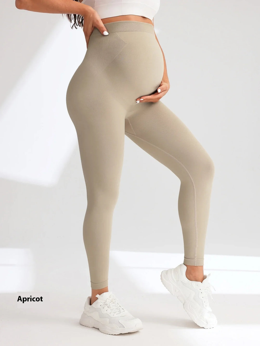 Pregnant woman wearing apricot maternity leggings and white sneakers, standing against white background
