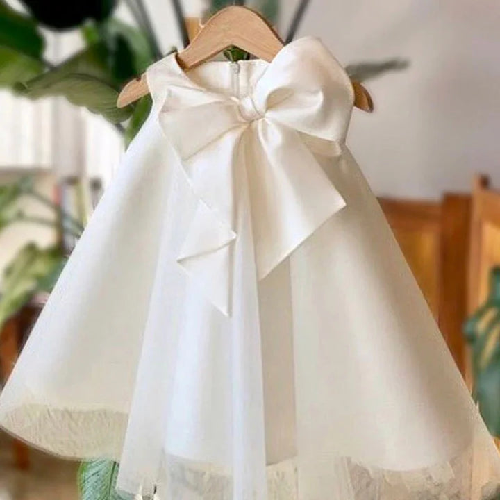 Elegant white baby dress with large satin bow hanging on wooden hanger indoors
