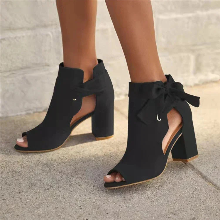 Black open-toe ankle boots with block heels and side cutouts, featuring a bow detail on outdoor pavement
