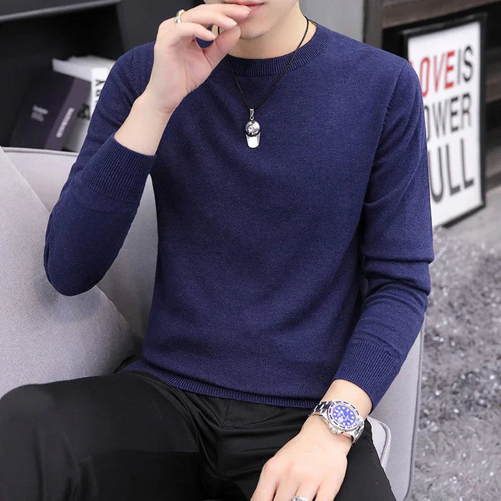 Man wearing a navy blue long-sleeve sweater with black pants, silver watch, and pendant necklace sitting on gray chair