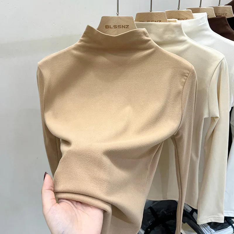Hand holding beige turtleneck top with other neutral-colored turtleneck tops on hangers in background