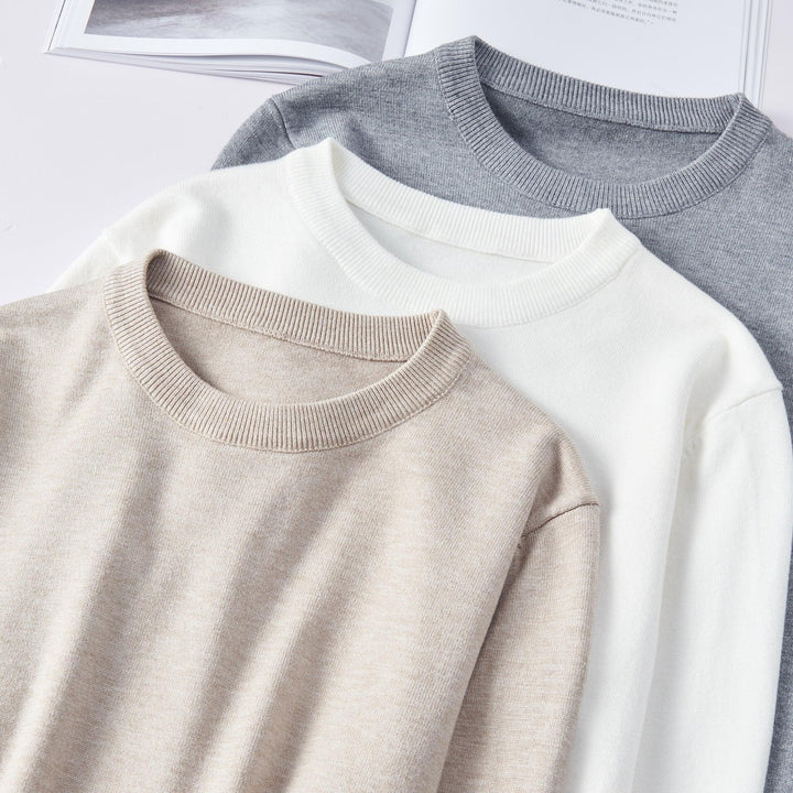 Three folded knit sweaters in beige, white, and gray, displayed on a white surface with an open book