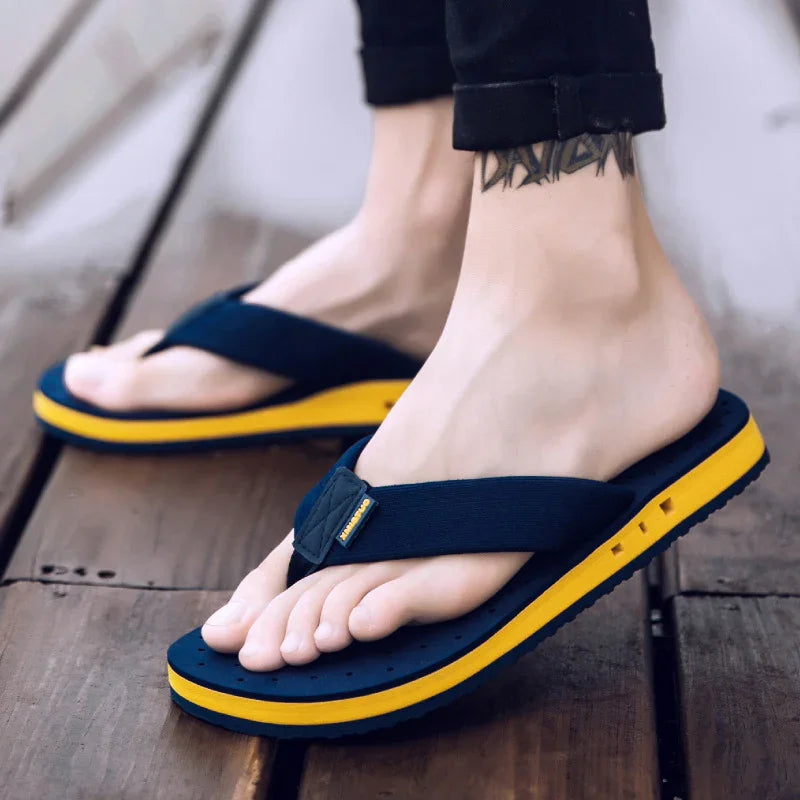 Close-up of feet wearing navy blue and yellow flip-flops on wooden floor, male tattoo visible