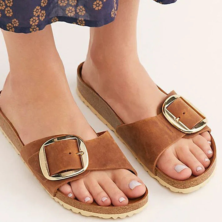 Close-up of feet wearing brown suede slide sandals with large gold buckle on cork sole