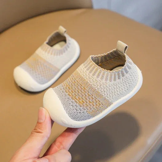 Light grey knitted baby shoes with white rubber soles on beige surface