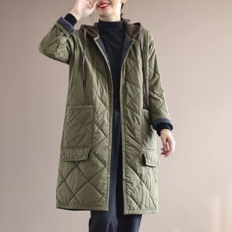 Woman wearing long olive green quilted coat with large front pockets and hood, black turtleneck and pants