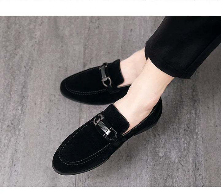 Black suede men's loafers with metal buckle detail worn with black pants on gray floor
