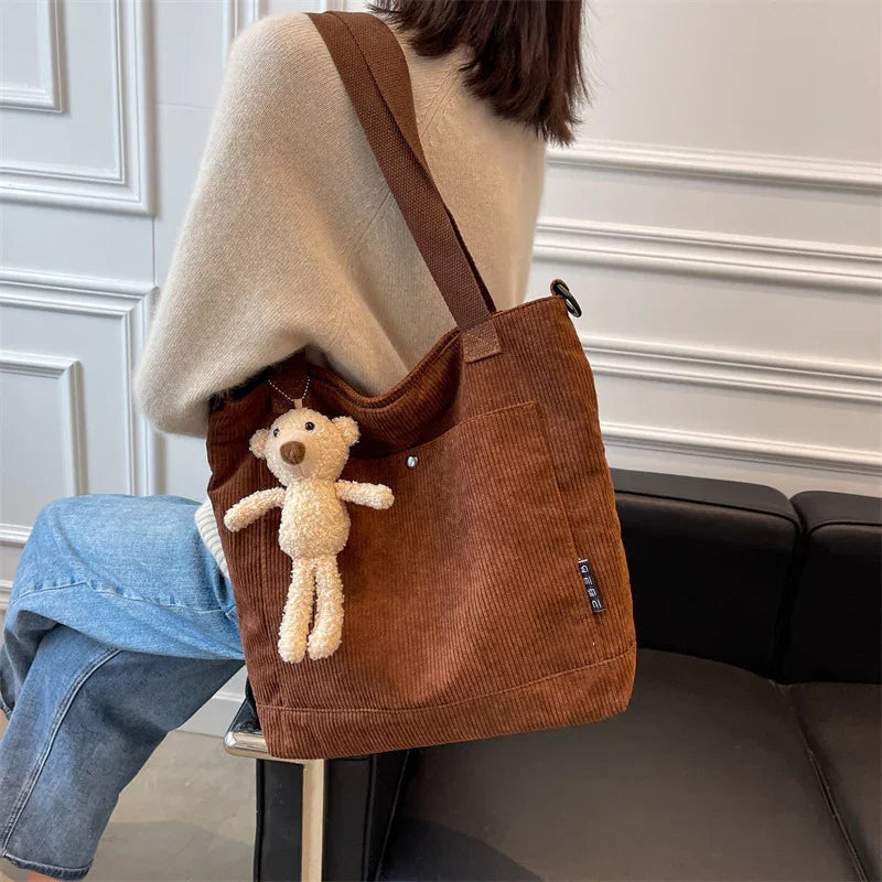 Brown corduroy shoulder bag with beige plush bear keychain worn by person in beige sweater and blue jeans