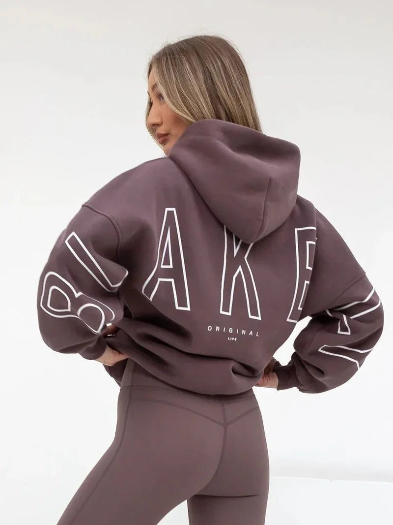 Woman wearing brown oversized hoodie with bold 'BLAKEN' text and fitted leggings in a studio