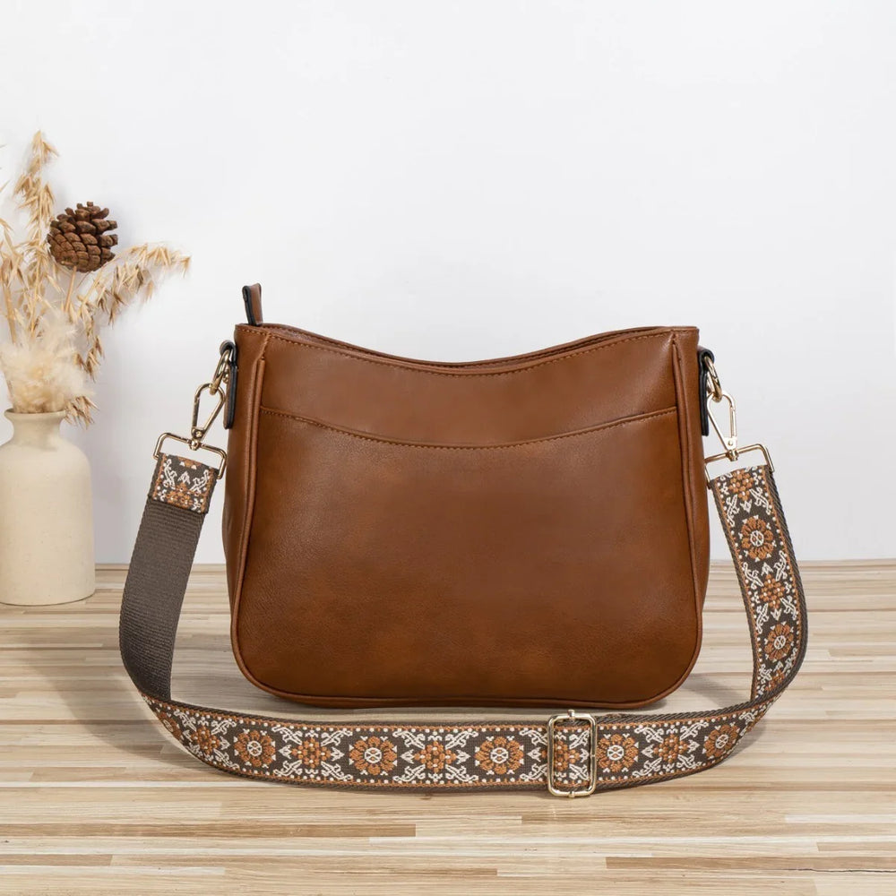Brown leather shoulder bag with patterned adjustable strap on wooden surface