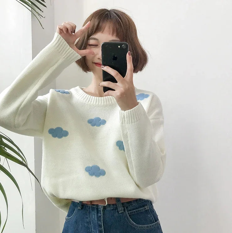 Woman taking selfie wearing white sweater with blue cloud patterns and blue jeans near green plant