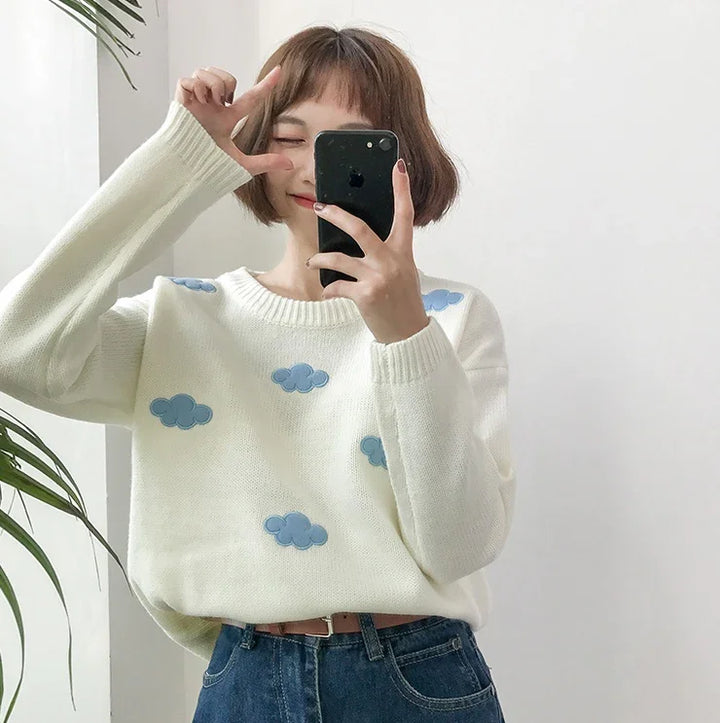 Woman taking selfie wearing white sweater with blue cloud patterns and blue jeans near green plant