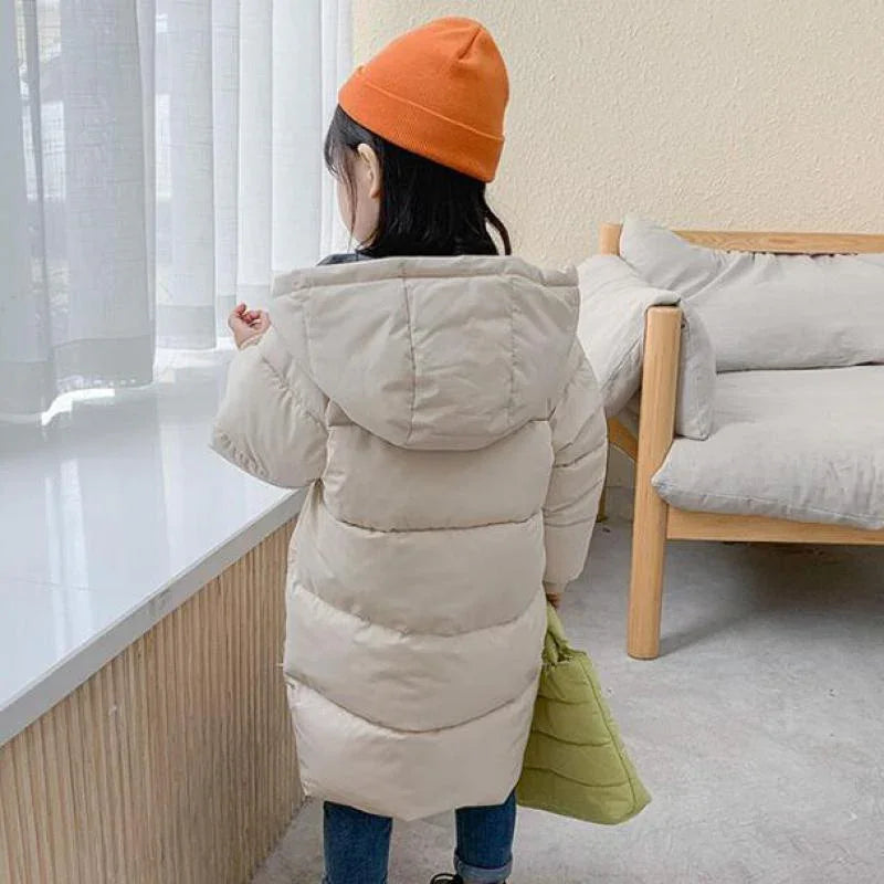 Child wearing beige puffer coat and orange beanie holding green bag indoors near a window