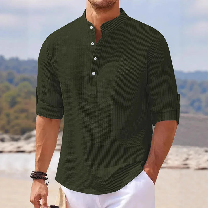 Man wearing dark green textured henley shirt with rolled sleeves and white pants at beach