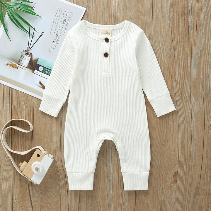 White ribbed long-sleeve baby romper with buttons on a wooden surface, styled with a toy camera and book