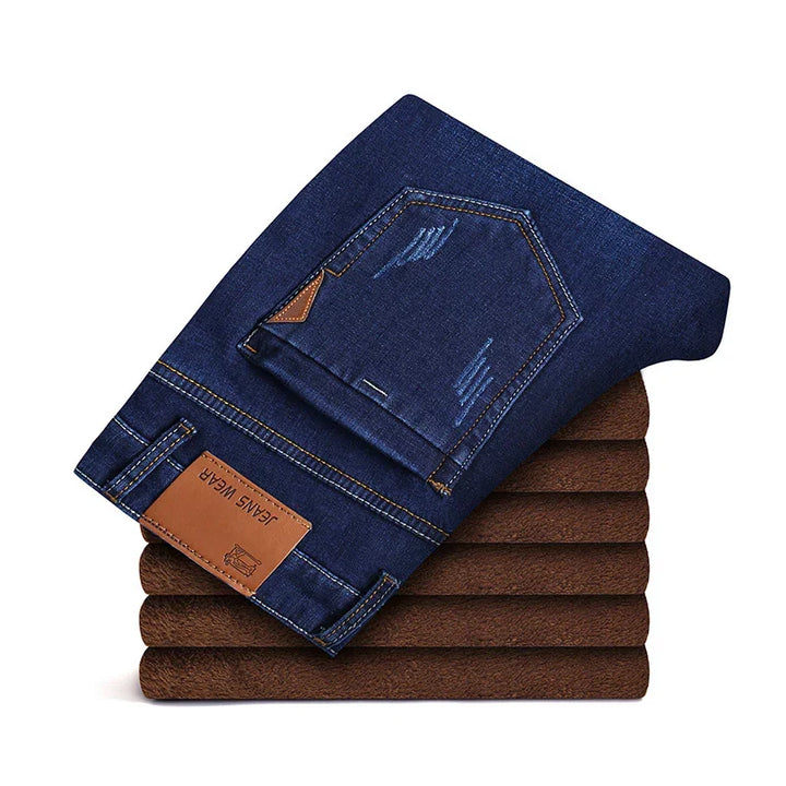 Folded dark blue denim jeans with brown leather patch labeled 'JEANS WEAR' stacked on brown fabric