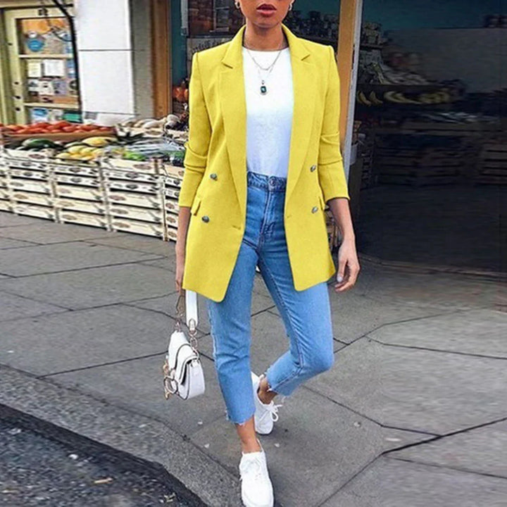 Woman wearing a yellow blazer, white top, blue jeans, and white sneakers walking on a city sidewalk