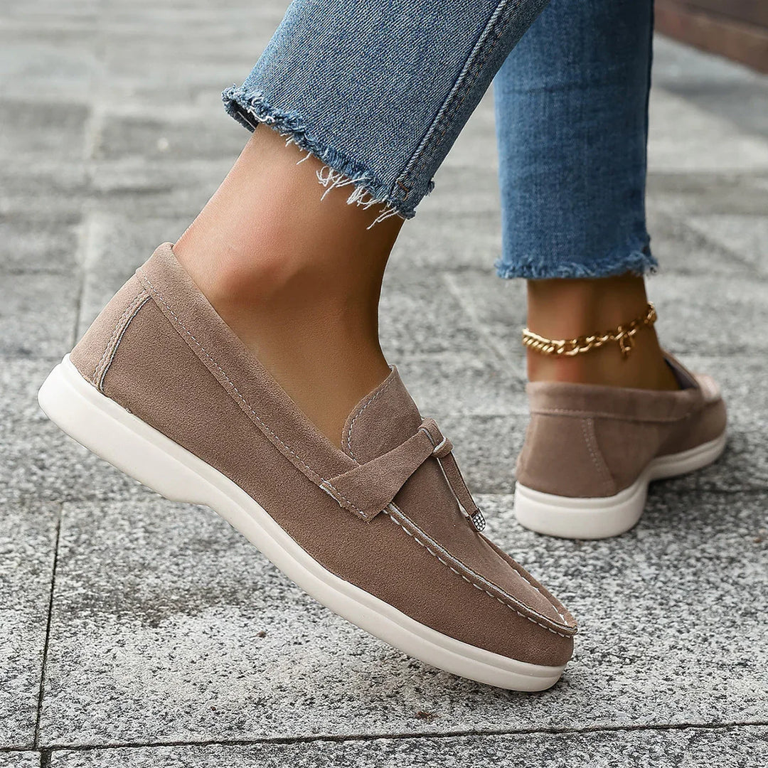 Close-up of suede beige loafers with white soles worn with frayed blue jeans and gold anklet on pavement