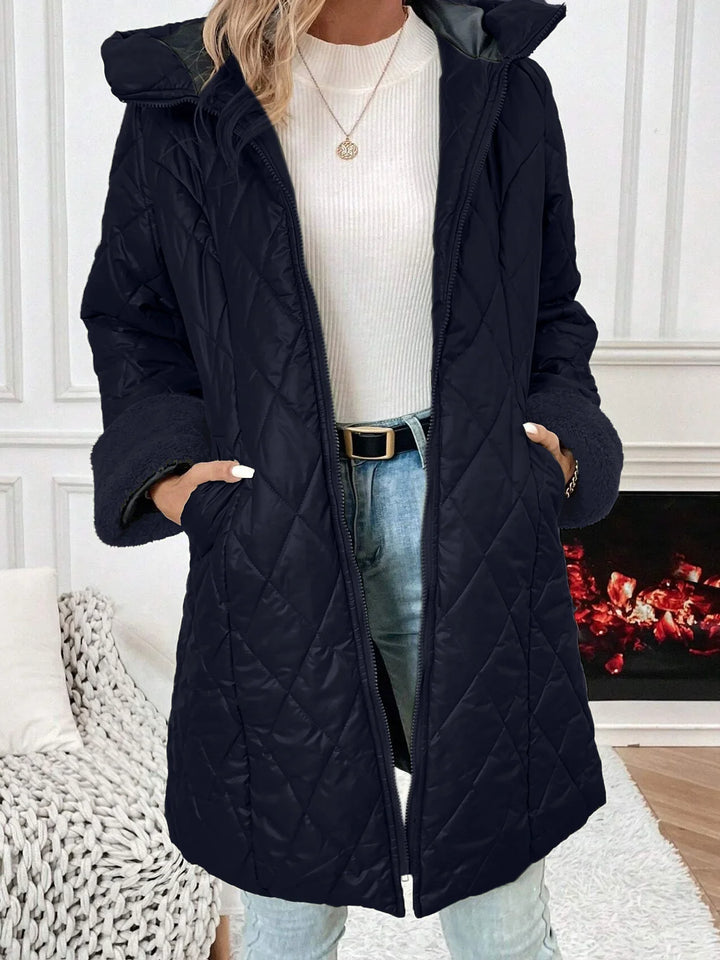 Woman wearing a black quilted winter coat with fleece cuffs over a white ribbed sweater and light blue jeans indoors