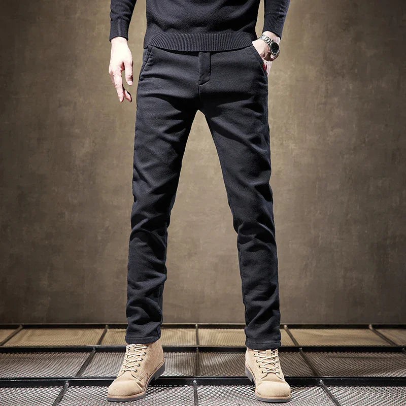 Man wearing slim black pants and beige suede boots standing against a brown textured backdrop
