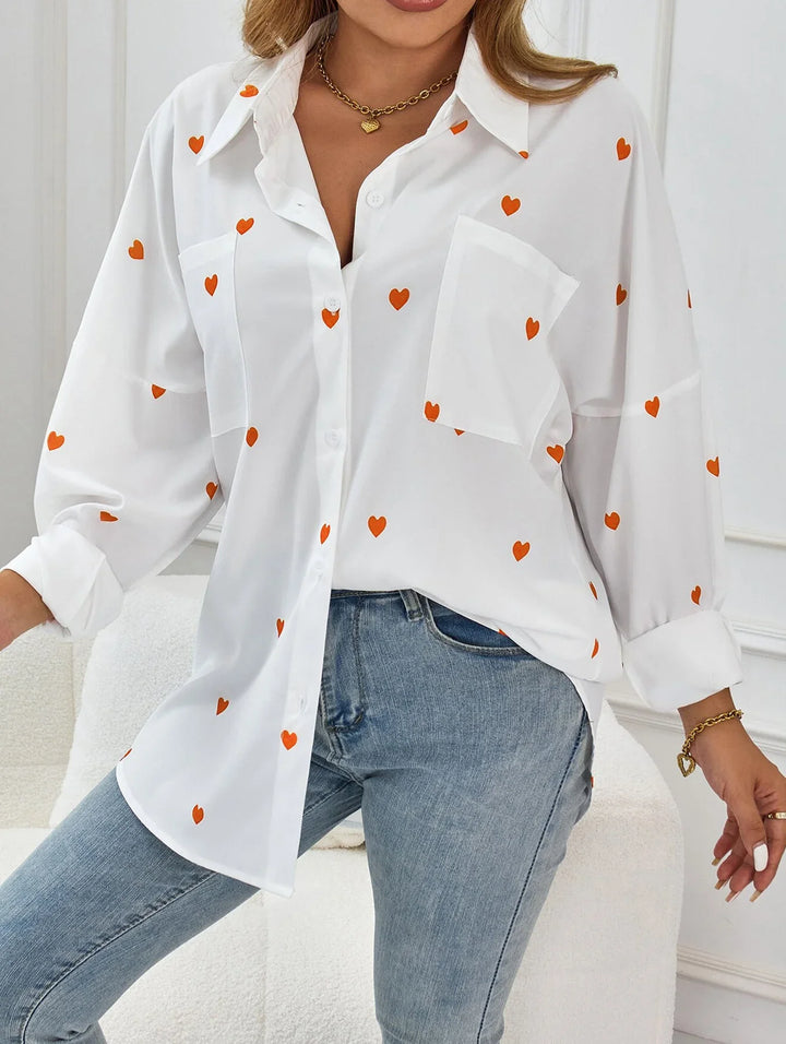 Woman wearing white button-up blouse with orange heart pattern and light blue jeans in indoor setting