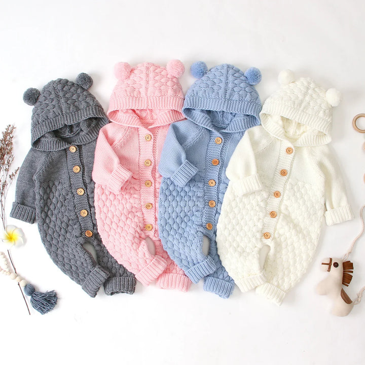Four knitted baby rompers in gray, pink, blue, and white with hoods and pom-poms, wooden buttons