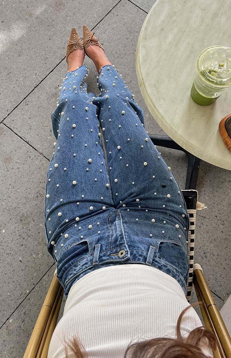 Person sitting wearing pearl-embellished blue jeans and brown pointed shoes next to table with green drink