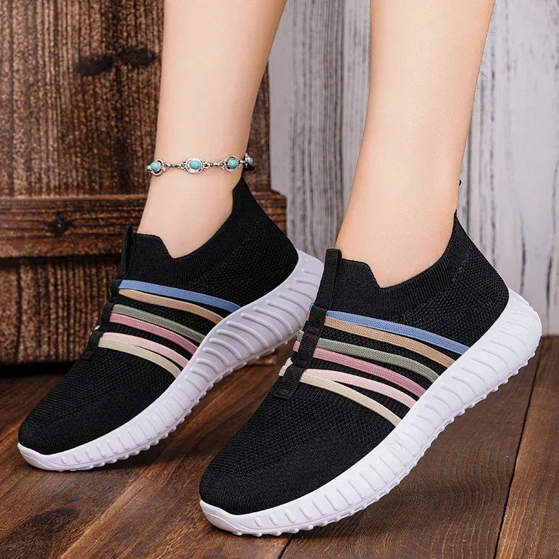 Black slip-on sneakers with white soles and multicolor elastic bands on wooden floor