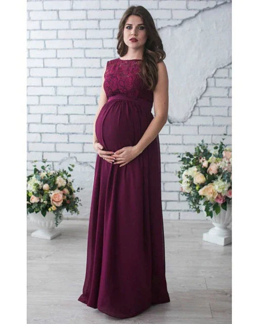 Pregnant woman in sleeveless burgundy lace maternity gown posing against white brick wall with floral arrangements