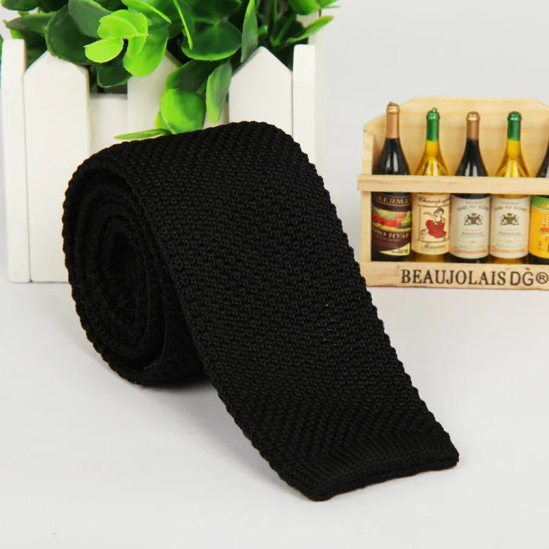Black knitted rolled necktie displayed with green plant and miniature wine bottles in wooden crate