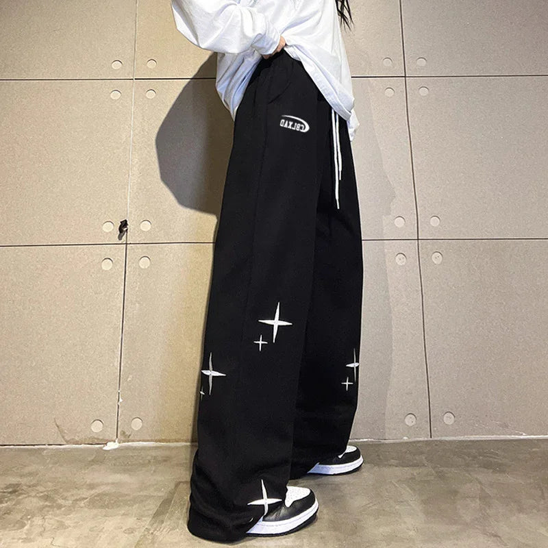 Person wearing black wide-leg sweatpants with white star designs and white drawstrings, paired with black and white sneakers