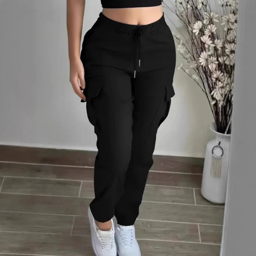 Woman wearing black cargo pants and white sneakers standing indoors near a vase with dried flowers