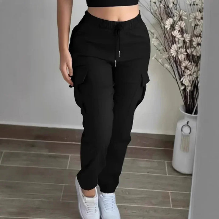 Woman wearing black cargo pants and white sneakers standing indoors near a vase with dried flowers