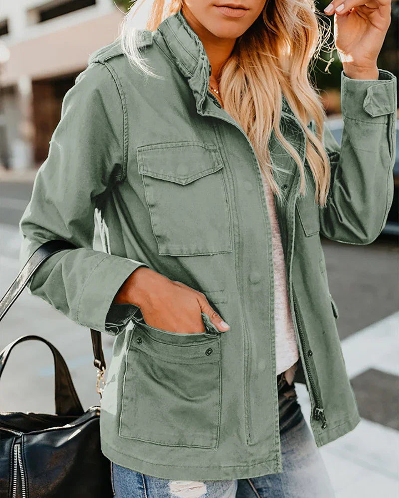 Woman wearing a green utility jacket with flap pockets and a zipper, posing outdoors holding a black bag