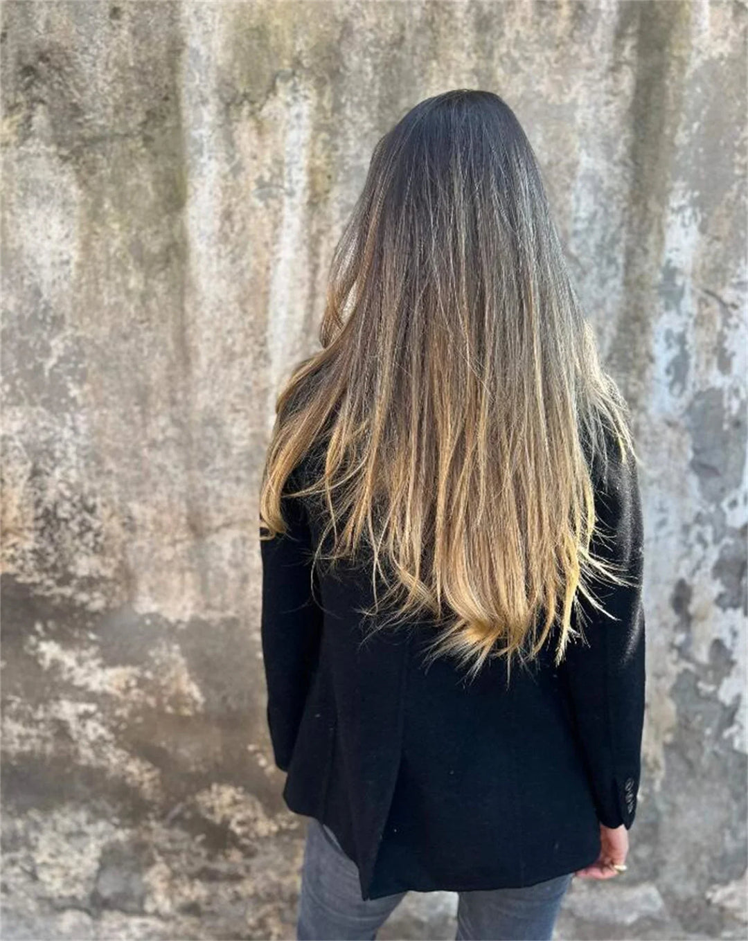Woman with long ombre hair wearing a black jacket standing against a textured concrete wall
