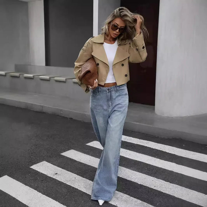 Woman in cropped beige trench coat, white crop top, wide-leg jeans, sunglasses, holding woven clutch on city street