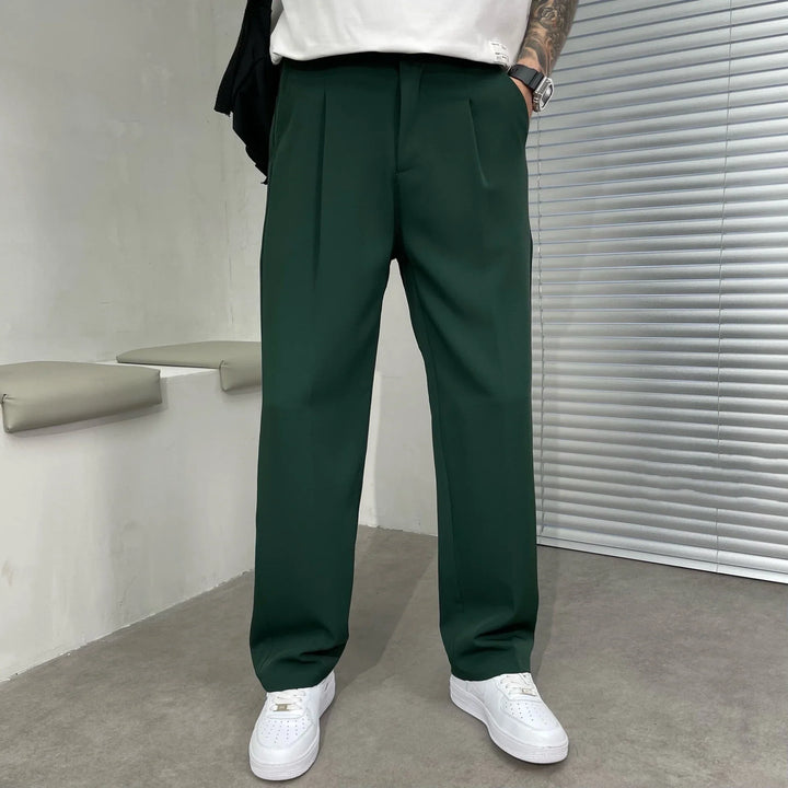 Man wearing dark green pleated trousers and white sneakers in a minimalist room with gray blinds